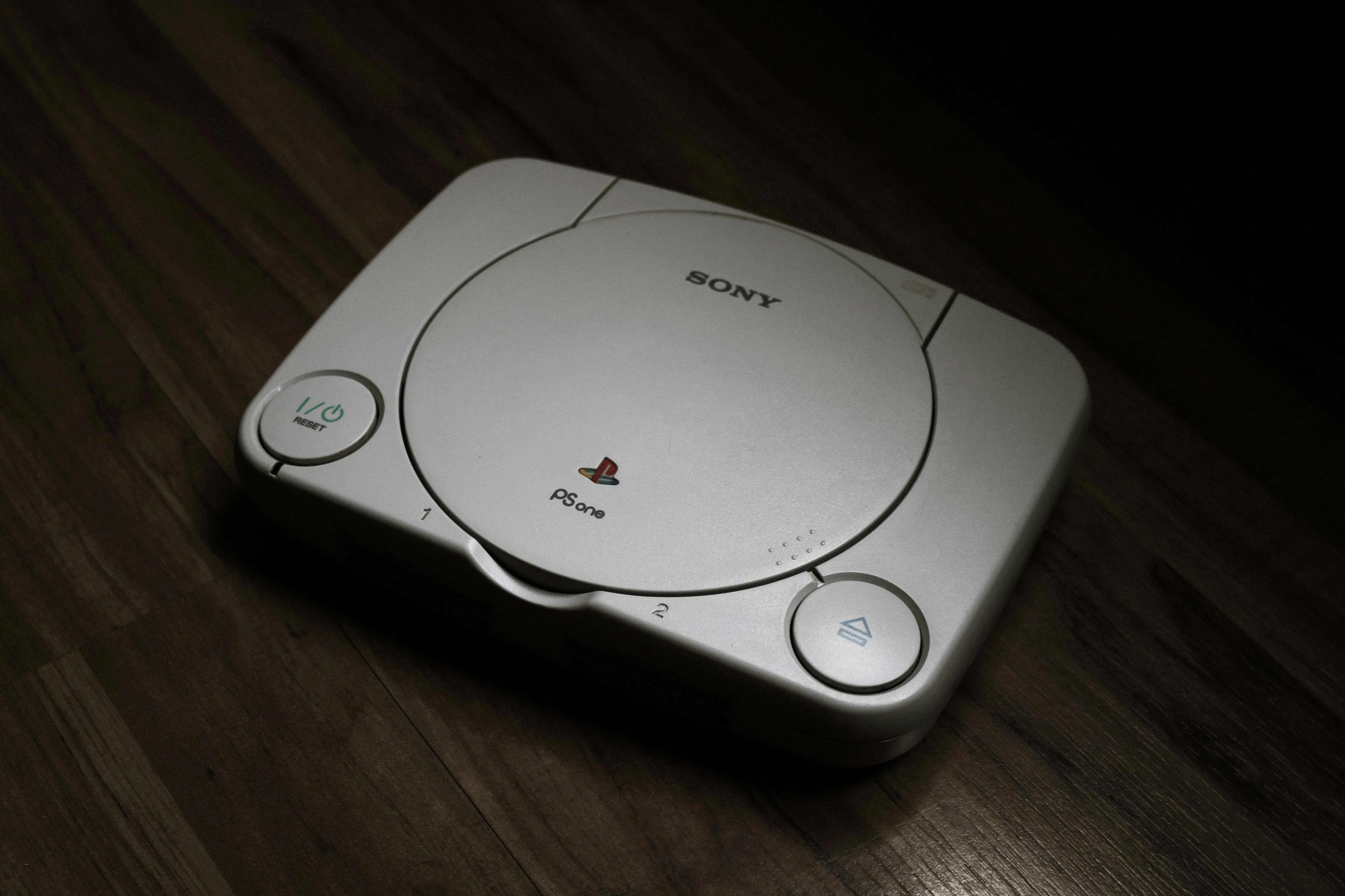 Máy console PlayStation 1 màu xám trên nền sàn gỗ, biểu tượng của kỷ nguyên gaming thập niên 90